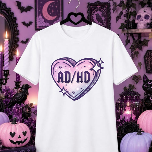 A white short-sleeve t-shirt on a black hanger, featuring a pastel pink and purple heart graphic with the text 'AD/HD' in the center. The shirt is displayed against a Halloween-themed purple background with pumpkins, candles, and black birds.