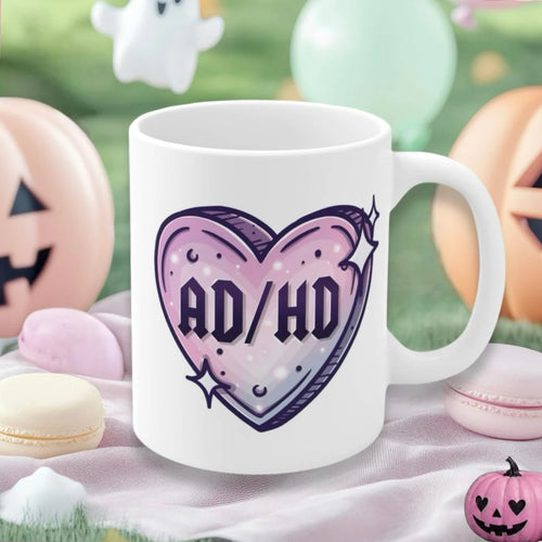 A white ceramic mug with a purple and pink heart graphic that reads 'AD/HD', styled on a soft pink cloth with Halloween-themed props like macarons, pumpkins, and a ghost in a blurred outdoor background.