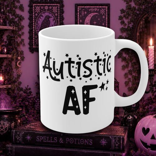 A white ceramic mug with the text 'Autistic AF' printed in black, styled on a dark book surrounded by purple and gothic Halloween decorations.