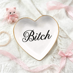 A white heart-shaped ceramic jewelry tray with a gold rim and the word 'Bitch' in black script, styled on a white lace fabric with pink ribbons, a pearl bracelet, and a small pink plush cat.