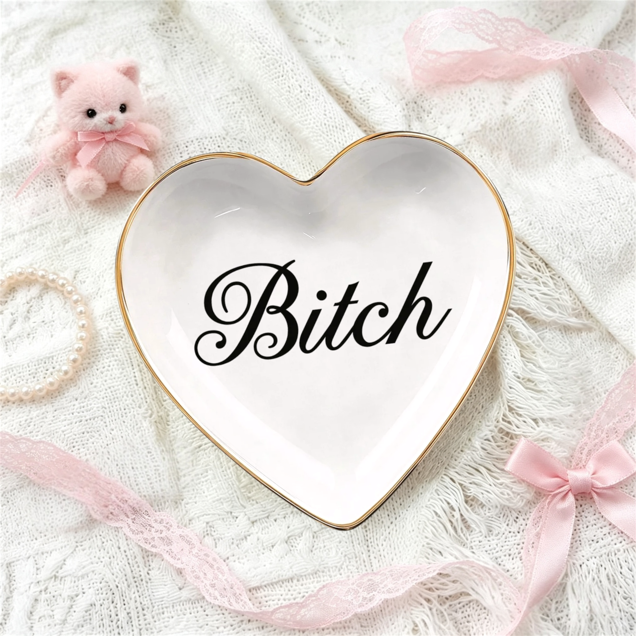 A white heart-shaped ceramic jewelry tray with a gold rim and the word 'Bitch' in black script, styled on a white lace fabric with pink ribbons, a pearl bracelet, and a small pink plush cat.