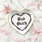 A heart-shaped ceramic je week tray with a gold rim and a black barbed wire graphic surrounding the gothic text 'Bad Bitch'. The tray is styled on a white lace fabric background with pink ribbons, a small pink plush toy, and a pearl necklace.
