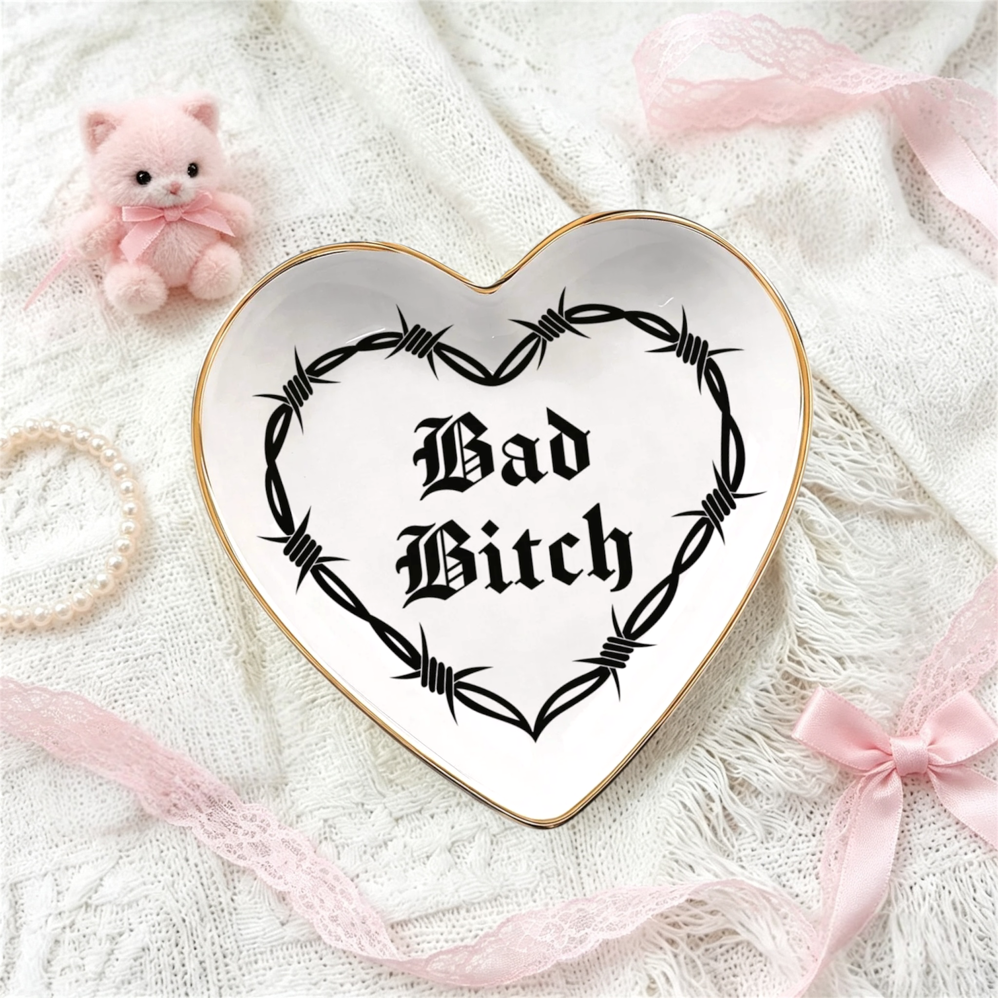 A heart-shaped ceramic je week tray with a gold rim and a black barbed wire graphic surrounding the gothic text 'Bad Bitch'. The tray is styled on a white lace fabric background with pink ribbons, a small pink plush toy, and a pearl necklace.