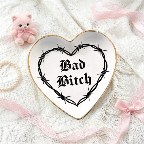 A heart-shaped ceramic je week tray with a gold rim and a black barbed wire graphic surrounding the gothic text 'Bad Bitch'. The tray is styled on a white lace fabric background with pink ribbons, a small pink plush toy, and a pearl necklace.