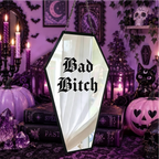 A coffin-shaped Halloween mirror with a black frame hanging in a spooky purple-themed room. The mirror reflects a white curtain, and the words 'Bad Bitch' are printed in black gothic font on the front.