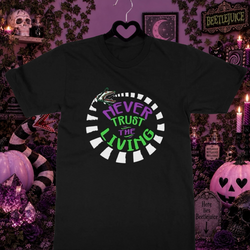 A black short-sleeve Beetlejuice t-shirt hanging on a black hanger, featuring a circular graphic with the text 'NEVER TRUST THE LIVING' in purple and green lettering, surrounded by white teeth-like shapes. The shirt is displayed against a highly decorated, spooky Halloween-themed background with pumpkins, candles, and purple walls.