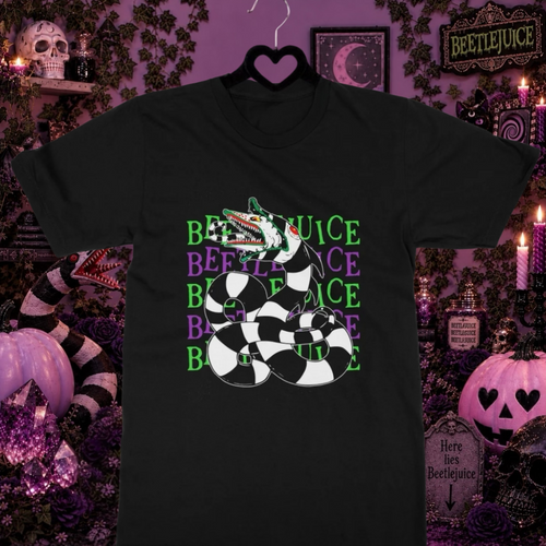 A black graphic Beetlejuice Sandworm t-shirt displayed on a black hanger against a highly detailed, spooky Halloween-themed background. The shirt features a graphic of a coiled black-and-white checkered snake with a green and white striped head, superimposed over the word 'BEETLEJUICE' repeated in green and purple.