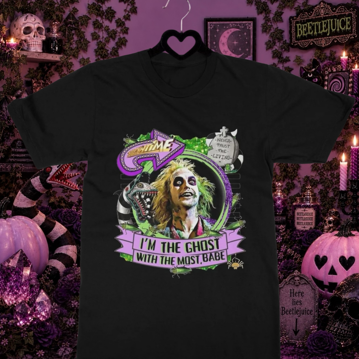 A black graphic t-shirt featuring Beetlejuice with the text 'I'M THE GHOST WITH THE MOST' displayed on a black hanger against a highly decorated, spooky Halloween-themed background with pumpkins, candles, and purple decor.
