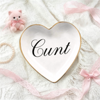 A heart-shaped white ceramic jewelry tray with a gold rim and the word 'C*nt' written in black script, styled on a white lace fabric with pink ribbons, a pearl bracelet, and a small pink plush bear.