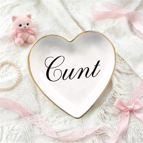 A heart-shaped white ceramic jewelry tray with a gold rim and the word 'C*nt' written in black script, styled on a white lace fabric with pink ribbons, a pearl bracelet, and a small pink plush bear.