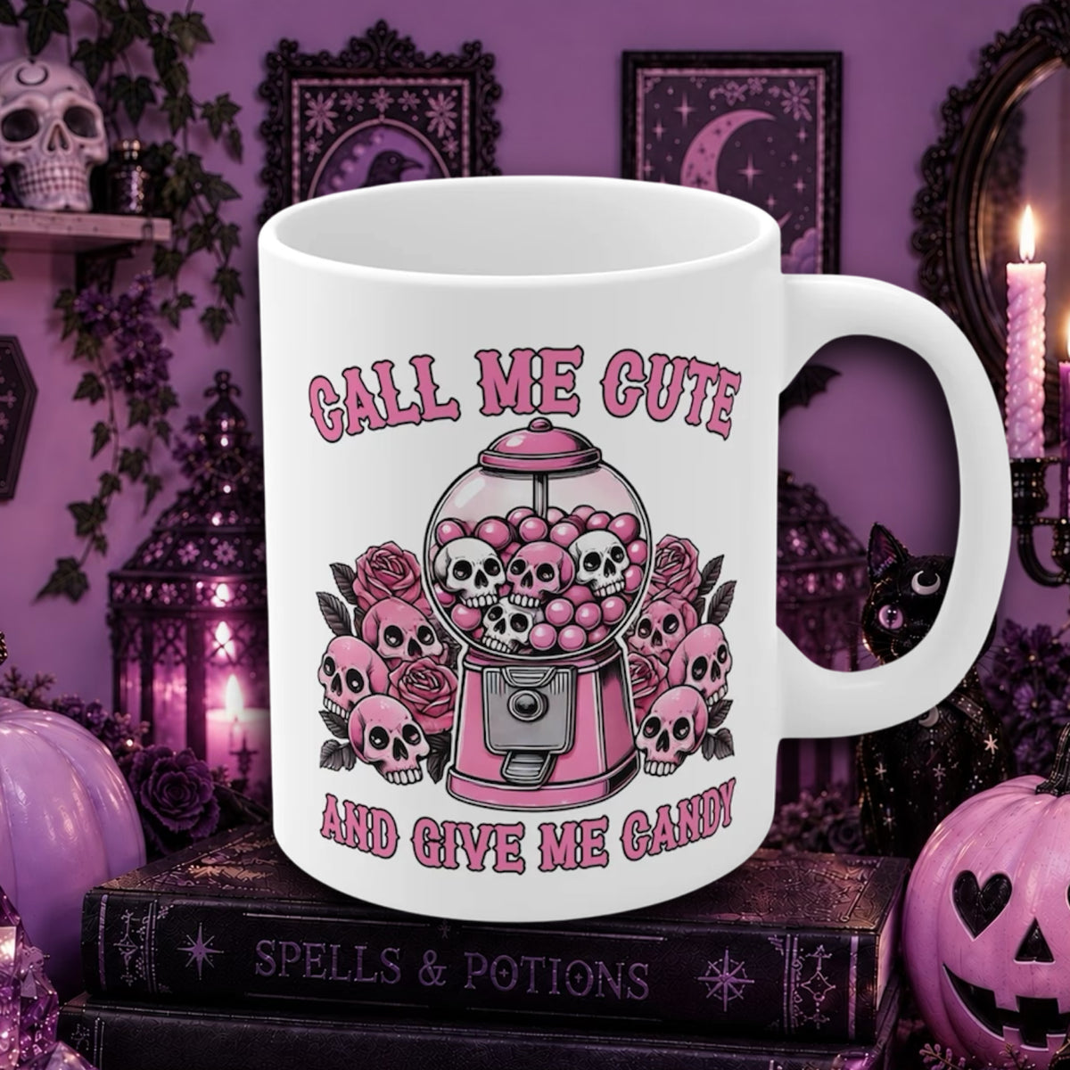 A white ceramic mug with a pink and black gothic graphic of a gumball machine filled with skulls and roses, surrounded by the text 'CALL ME CUTE AND GIVE ME CANDY'. The mug is displayed on a stack of books in a purple, Halloween-themed setting with candles, pumpkins, and framed art.