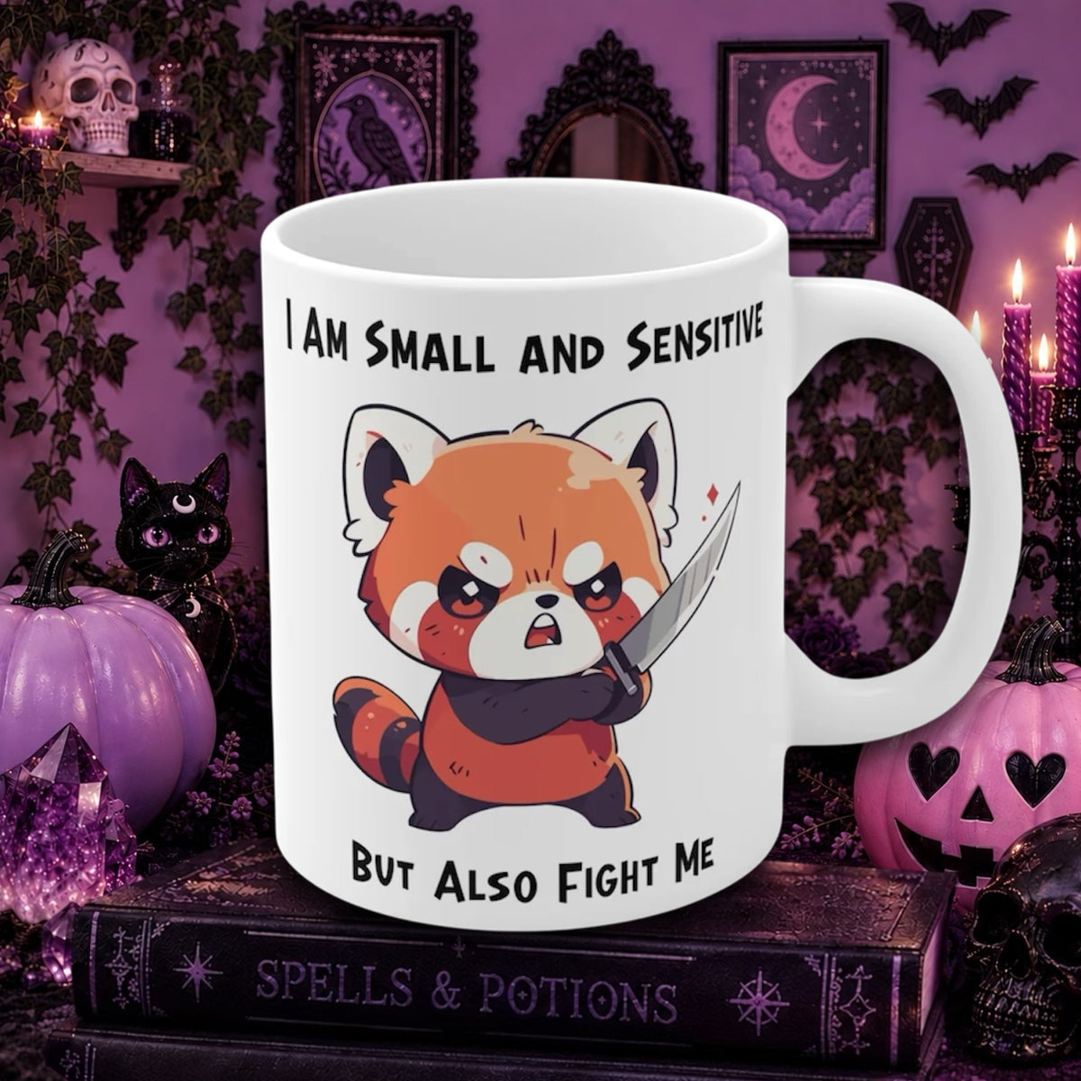 A white ceramic mug with a cartoon red panda holding a knife, surrounded by the text 'I AM SMALL AND SENSITIVE BUT ALSO FIGHT ME'. The mug is displayed on a stack of books in a spooky, Halloween-themed room with purple walls, pumpkins, and candles.