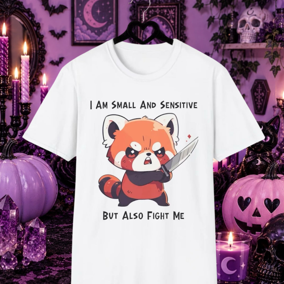 A white short-sleeve graphic T-shirt with a red panda holding a knife with the text 'I AM SMALL AND SENSITIVE BUT ALSO FIGHT ME'. Purple Halloween-themed background with pumpkins, candles, and gothic decor. 
