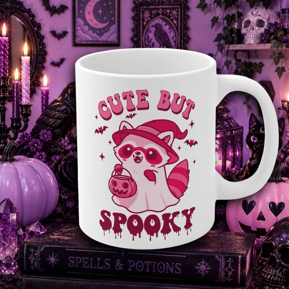 A white ceramic mug with a pink and red cartoon raccoon wearing a witch hat and holding a pumpkin, surrounded by the text 'CUTE BUT SPOOKY'. The mug is set against a dark, gothic Halloween-themed background with purple candles, pumpkins, and books.