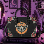 A black tote bag with gothic Halloween-themed artwork, including a central moth-and-skull motif, roses, and decorative corners, displayed on a book in a purple witch-themed room.