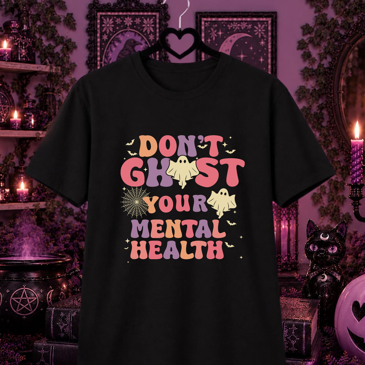 A black graphic t-shirt hanging on a hanger against a dark, gothic Halloween-themed background. The shirt features a colorful graphic that reads 'DON'T GHOST YOUR MENTAL HEALTH' in pastel pink, orange, and purple lettering, decorated with cartoon ghosts, bats, and a spiderweb.