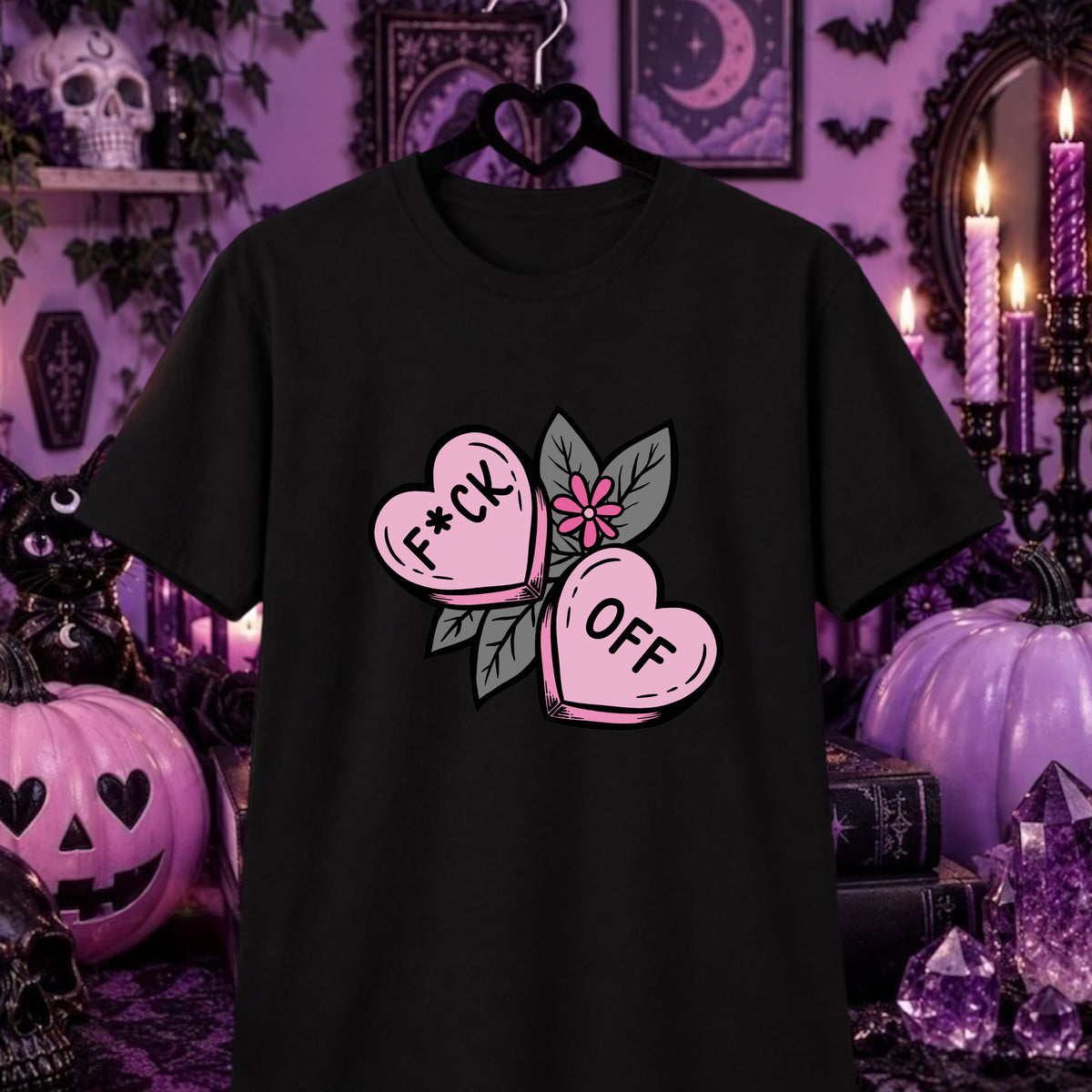 A black short-sleeve t-shirt hanging on a black heart-shaped hanger against a purple Halloween-themed background. The shirt features a graphic of two pink candy hearts that spell out 'F*CK OFF' with green leaves and a small pink flower in the center.
