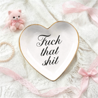 A heart-shaped white ceramic jewelry tray with a gold rim and the phrase 'Fuck that shit' in black script, styled on a white knit blanket with pink ribbons, a small pink plush bear, and a pearl bracelet.