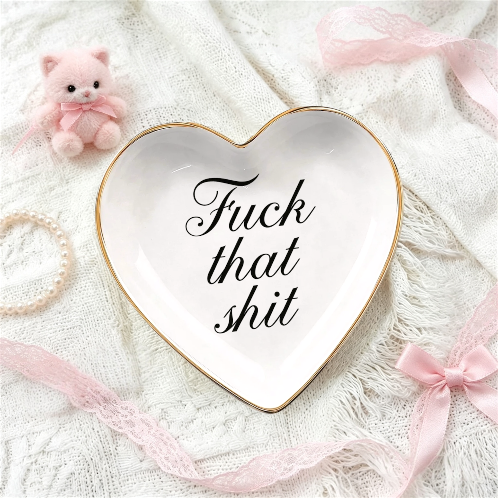 A heart-shaped white ceramic jewelry tray with a gold rim and the phrase 'Fuck that shit' in black script, styled on a white knit blanket with pink ribbons, a small pink plush bear, and a pearl bracelet.
