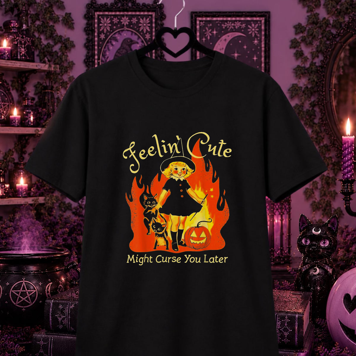 A black short-sleeve graphic t-shirt hanging on a black hanger against a dark, purple-lit Halloween-themed background. The graphic features a cartoon witch in a black dress standing in orange and yellow flames, with a black cat, a jack-o'-lantern, and the text 'Feelin' Cute' above and 'Might Curse You Later' below.