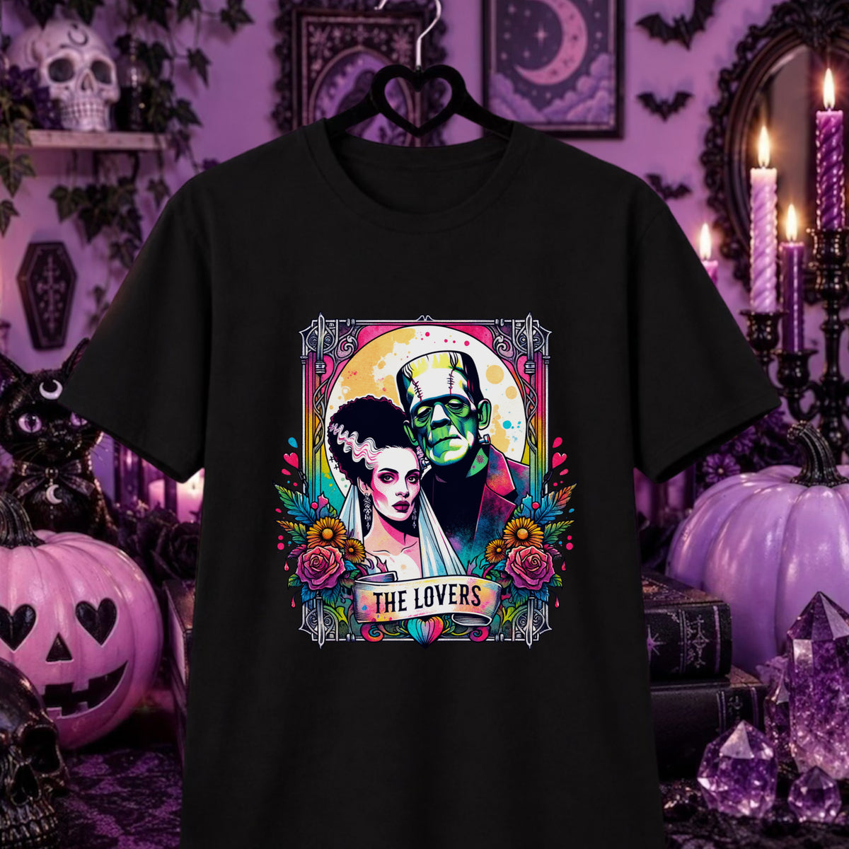 A black short-sleeve graphic t-shirt hanging on a black hanger against a Halloween-themed purple background. The graphic features a colorful Tarot illustration of Frankenstein's monster and Bride of Frankenstein with the text 'THE LOVERS' at the bottom.