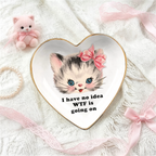 A heart-shaped white ceramic jewelry tray with a gold rim, featuring a cartoon kitten illustration and the text 'I have no idea WTF is going on'. The tray is styled on a white lace fabric with pink ribbons, a small pink plush kitten, and a pearl necklace.