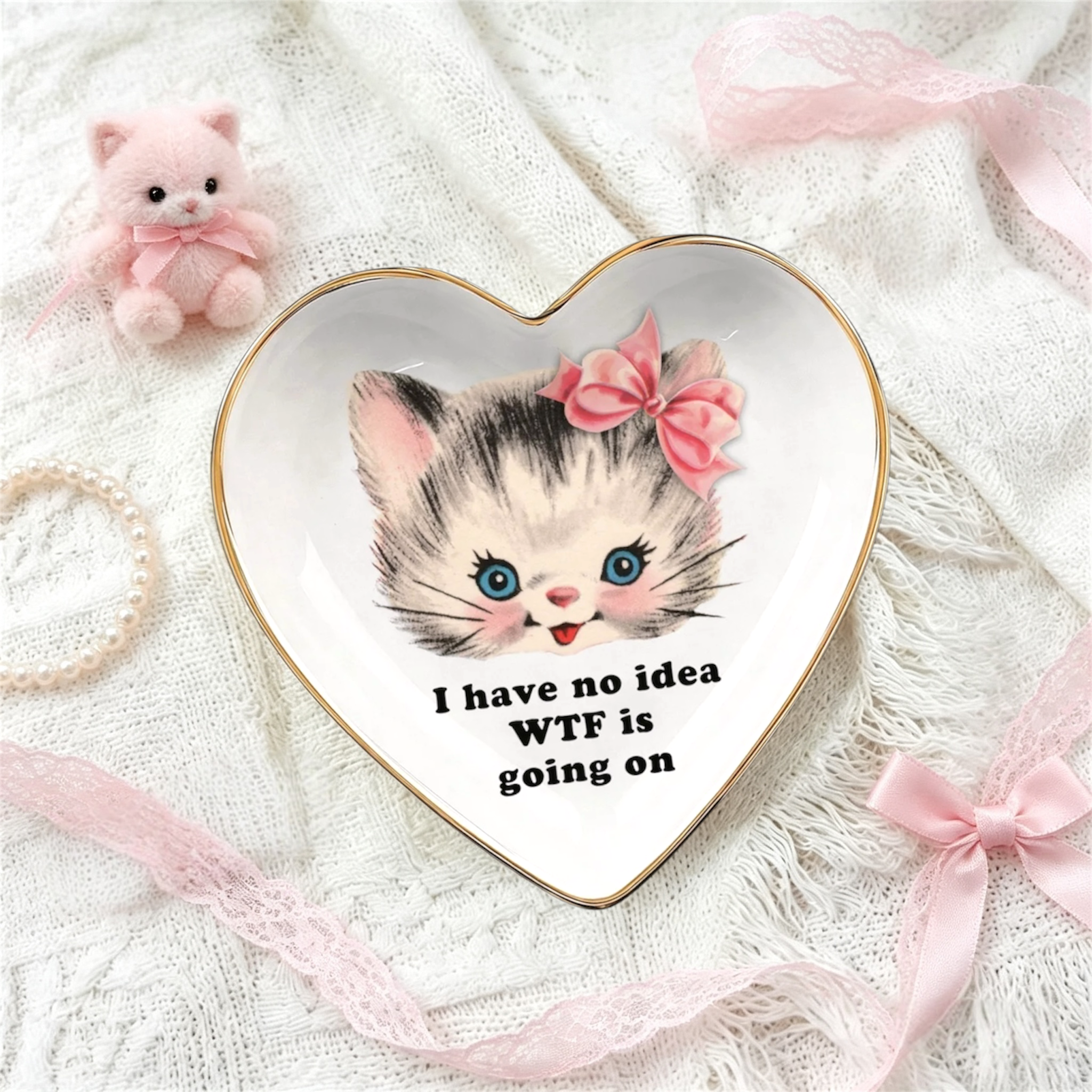 A heart-shaped white ceramic jewelry tray with a gold rim, featuring a cartoon kitten illustration and the text 'I have no idea WTF is going on'. The tray is styled on a white lace fabric with pink ribbons, a small pink plush kitten, and a pearl necklace.