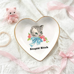 A heart-shaped ceramic jewelry tray with a gold rim, featuring a vintage-style illustration of a gray kitten with a blue bow and the text 'Stupid Bitch'. The tray is styled on a white lace fabric background with pink ribbons, a small pink plush cat, and a pearl necklace.