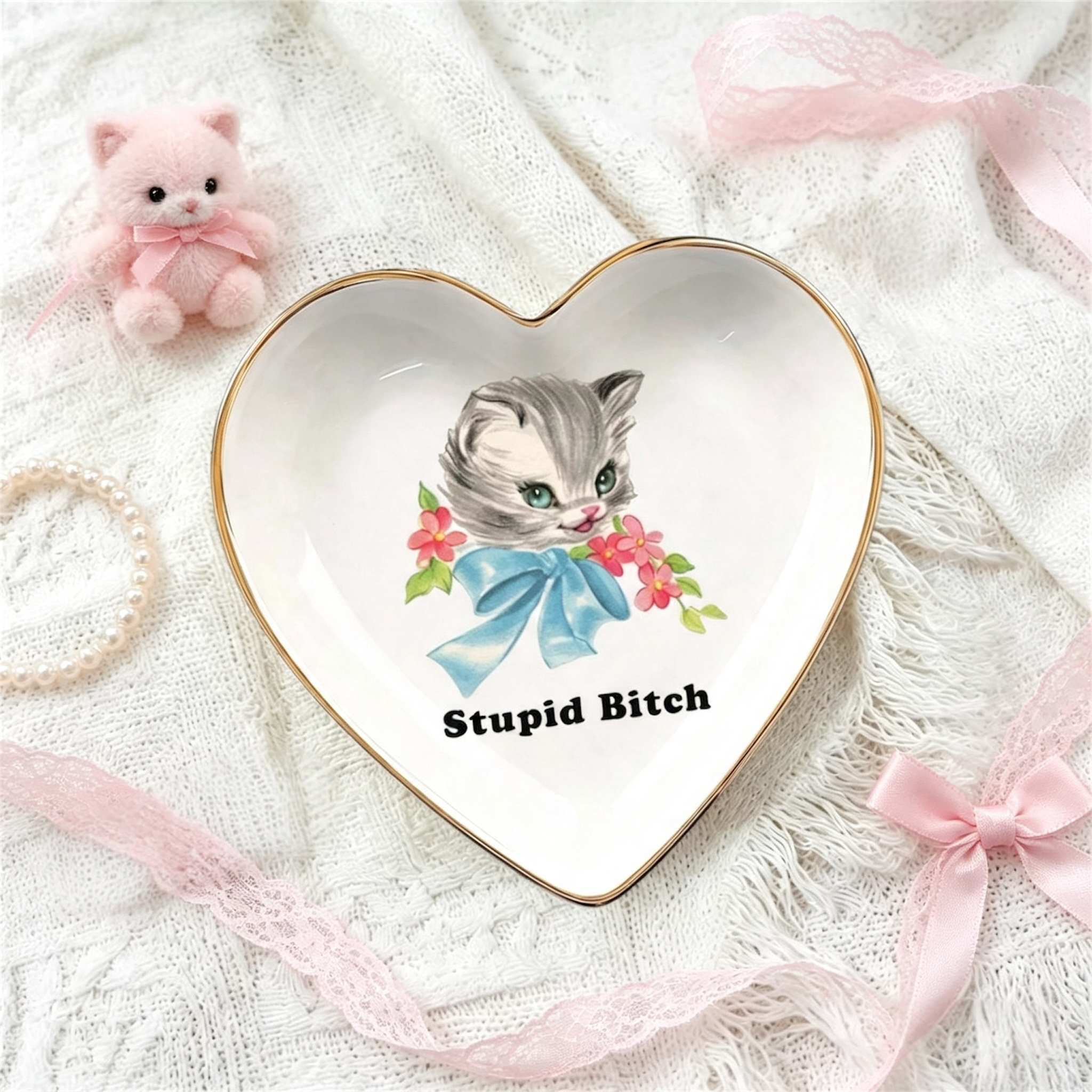 A heart-shaped ceramic jewelry tray with a gold rim, featuring a vintage-style illustration of a gray kitten with a blue bow and the text 'Stupid Bitch'. The tray is styled on a white lace fabric background with pink ribbons, a small pink plush cat, and a pearl necklace.