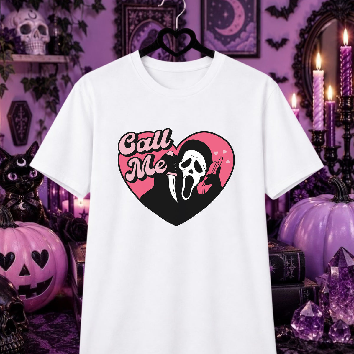 A white short-sleeve graphic T-shirt hanging on a black hanger against a Halloween-themed purple background with pumpkins, candles, and spooky decor. The shirt features a heart-shaped graphic with the words 'Call Me' in pink script over a black and pink heart containing a white Ghostface-style mask and a pink lipstick.
