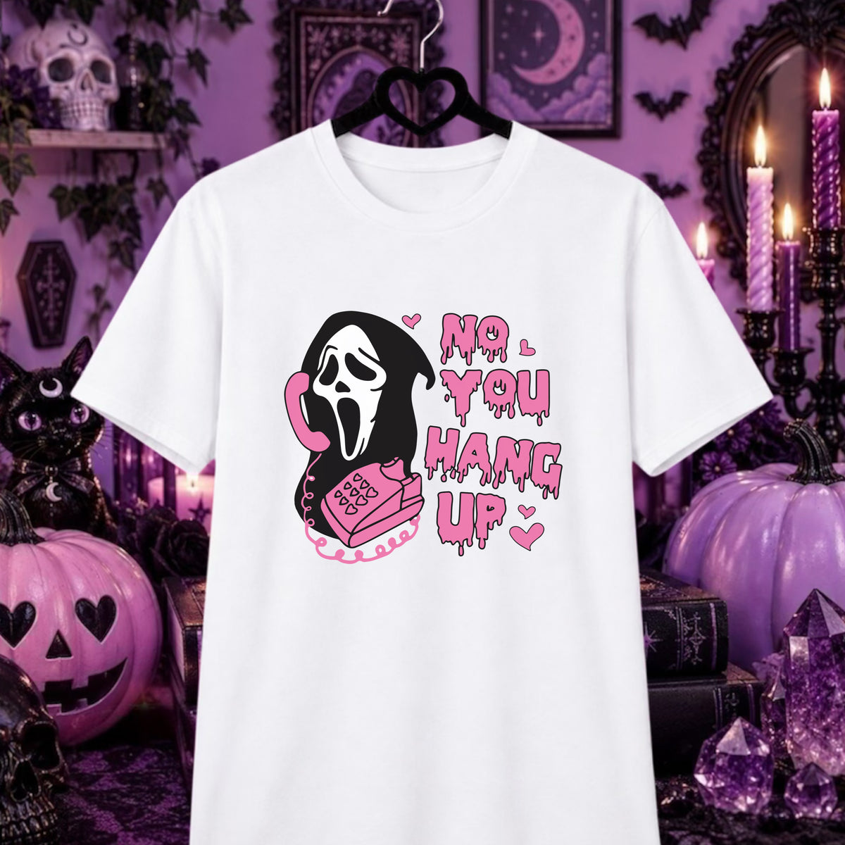 Ghostface no you hang up tshirt : A white short-sleeve graphic T-shirt hanging on a black hanger against a Halloween-themed purple background with pumpkins, candles, and gothic decor. The shirt features a Ghostface-style ghost character on a pink phone next to dripping pink text that reads 'NO YOU HANG UP'.
