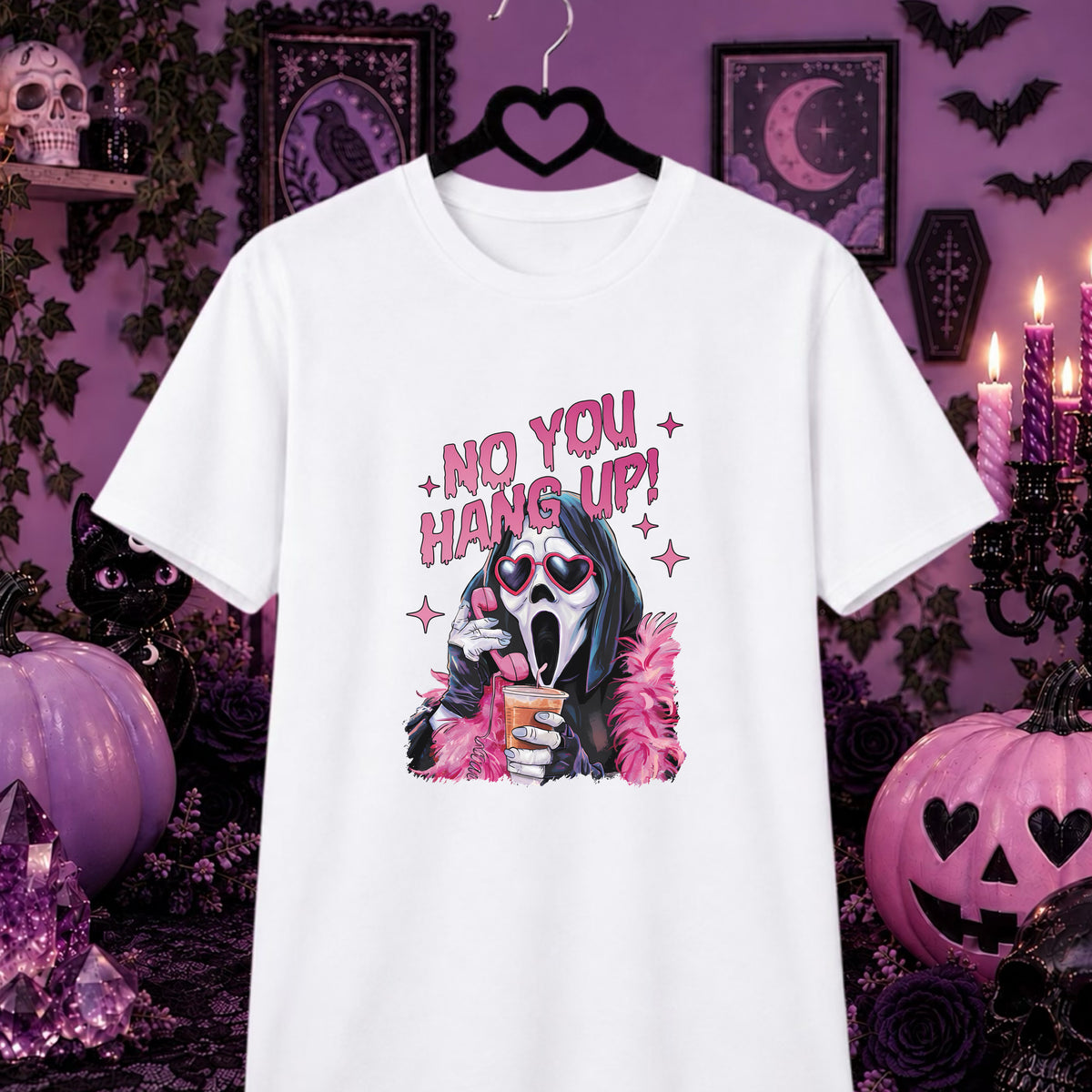 A white short-sleeve graphic T-shirt hanging on a black hanger against a Halloween-themed purple background with pumpkins, candles, and spooky decor. The shirt features a Ghostface-style illustration with pink text reading 'NO YOU HANG UP!' and pink feather accents.