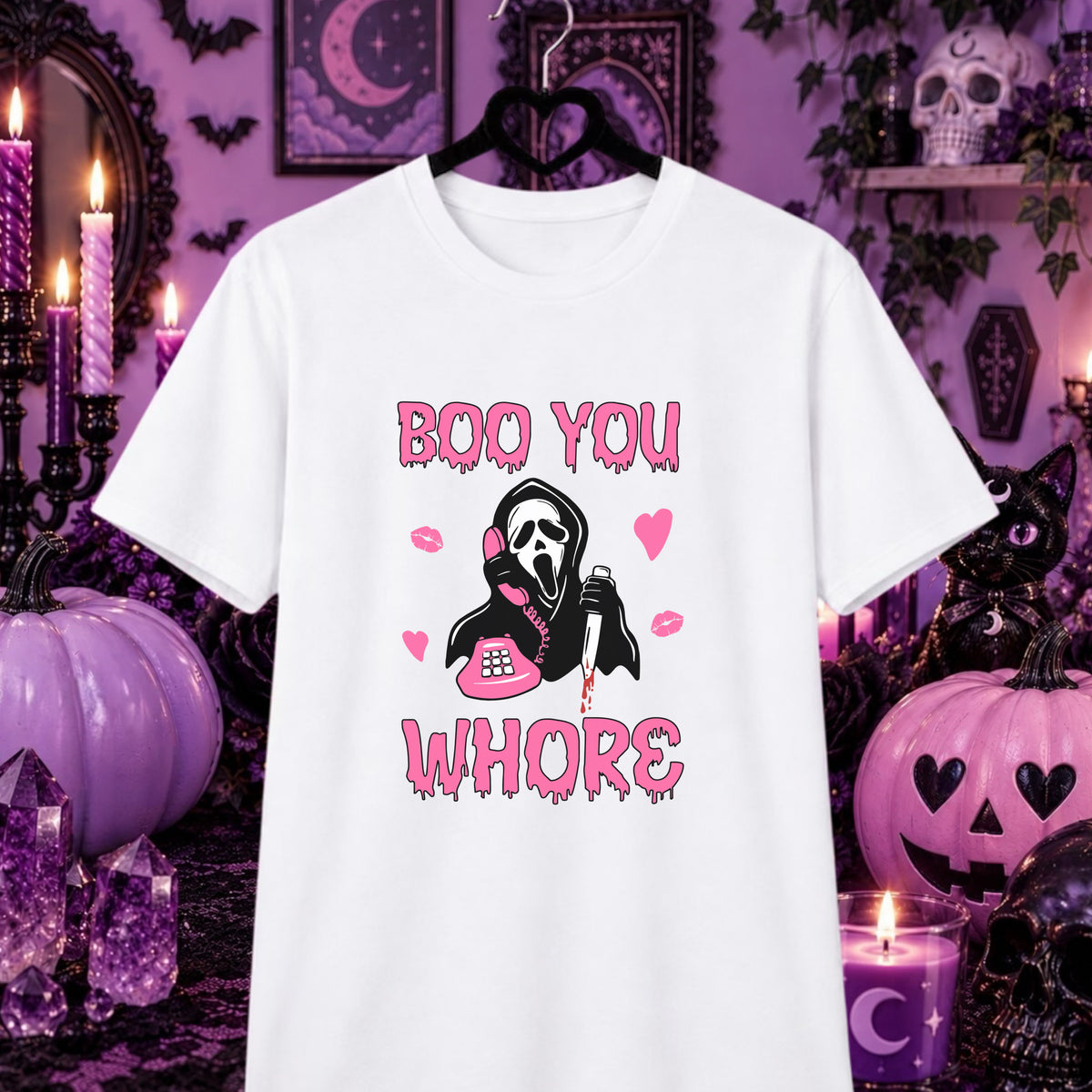 A white short-sleeve graphic t-shirt hanging on a black hanger against a Halloween-themed purple background. The graphic features the text 'BOO YOU WHOR' in a dripping pink font, surrounding a grim reaper illustration holding a pink phone and lipstick.