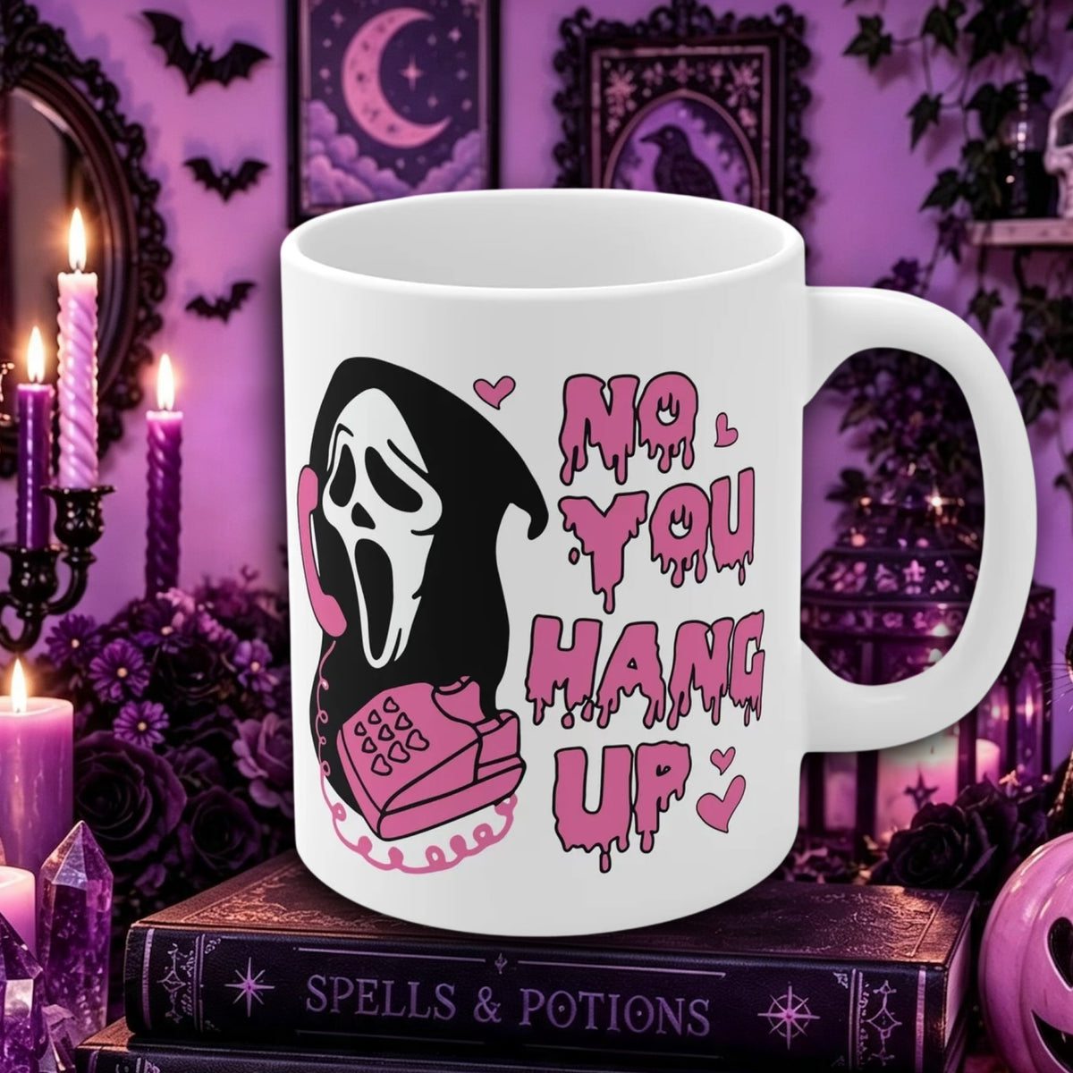 Ghostface no you hang up mug : A white ceramic mug with a gothic horror graphic of Ghostface on a pink phone and the text 'NO YOU HANG UP' in dripping pink letters, displayed on a stack of books in a purple, witch-themed setting with candles and crystals.