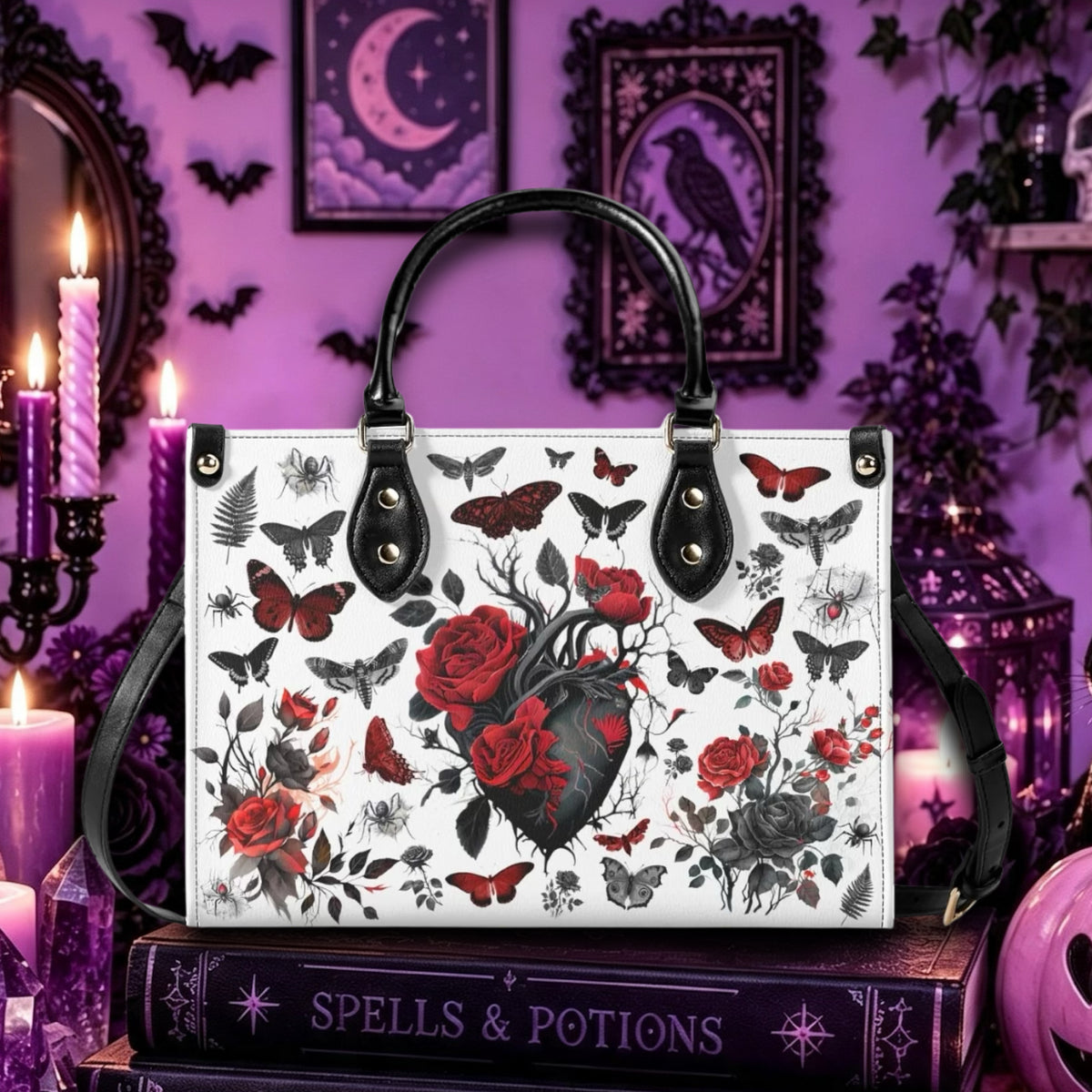 A white tote bag with black handles and a black-and-red gothic illustration print featuring an anatomical heart, roses, butterflies, and ravens, displayed on a purple gothic-themed background with candles and books.