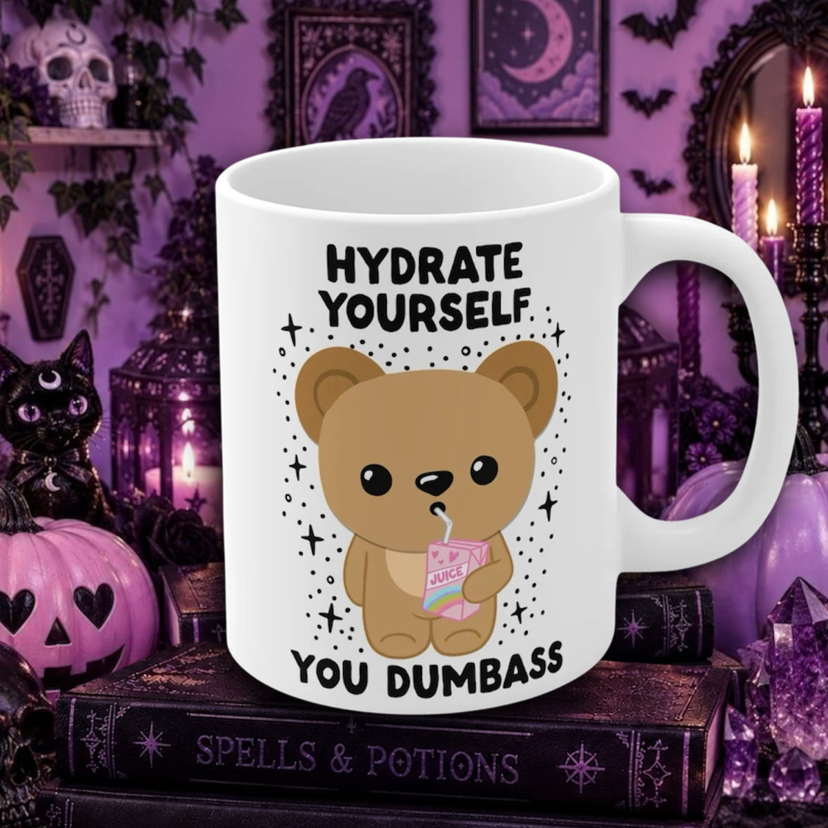 A white ceramic mug with a cartoon teddy bear holding a pink drink and the text 'HYDRATE YOURSELF YOU DUMBSS' printed on it, displayed on a stack of gothic books in a Halloween-themed purple room with candles, a black cat, and a pumpkin.