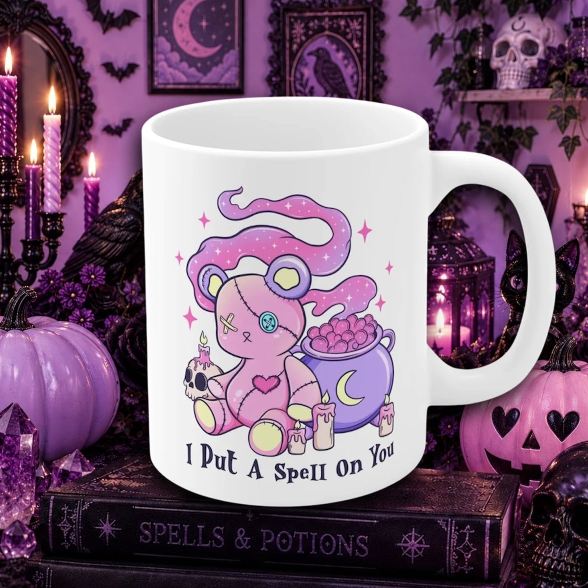 A white ceramic mug with a pastel goth teddy bear casting a spell next to a purple cauldron, with the text 'I Put A Spell On You' printed below. The mug is displayed on a gothic-themed book surrounded by purple pumpkins, candles, and witch hats.