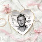A heart-shaped ceramic jewelry tray with a gold rim, featuring a black-and-white mugshot-style portrait on a height chart background. The tray is styled on a white textured knit blanket with pink ribbons, a pearl bracelet, and a small pink plush toy nearby.