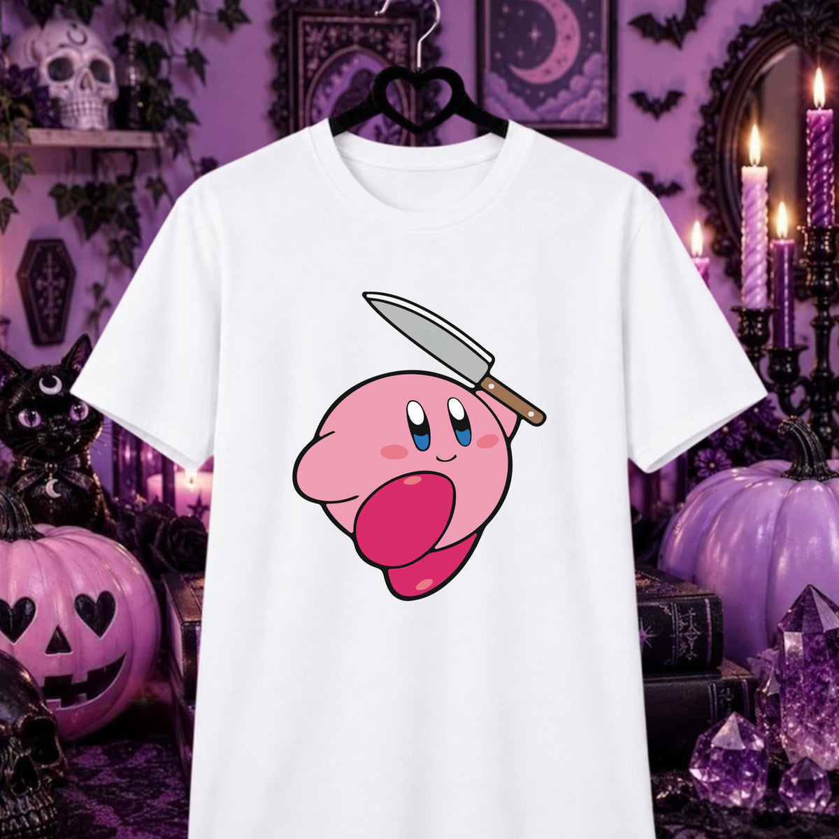 A white short-sleeve t-shirt hanging on a black hanger, featuring a cartoon graphic of Kirby with a knife on its head. The background is a Halloween-themed setting with purple candles, pumpkins, and spooky decor.