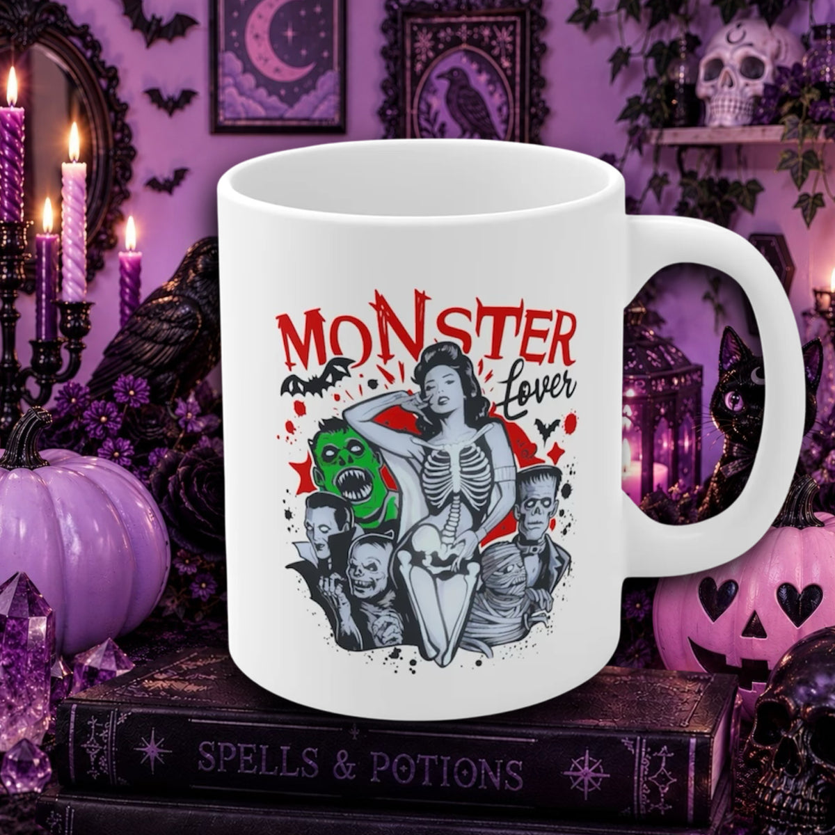 A white ceramic mug with a 'Monster Lover' graphic print, styled on a dark book surrounded by Halloween decorations like purple pumpkins, candles, and a black cat in a purple gothic room.