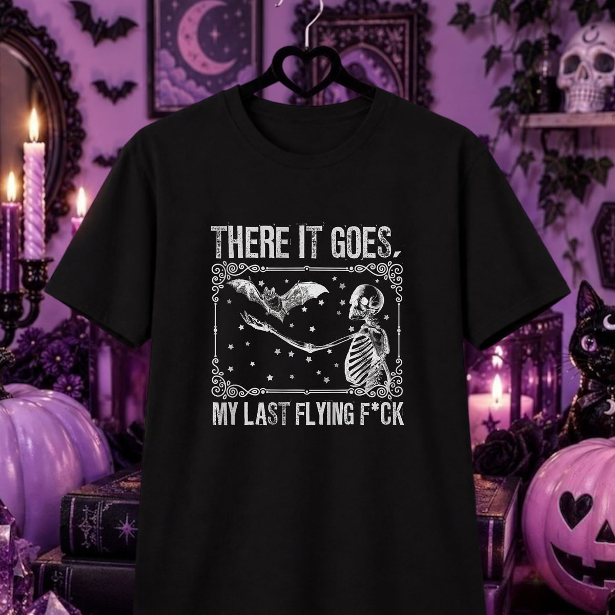 A black short-sleeve graphic t-shirt hanging on a black hanger against a purple Halloween-themed background with candles, pumpkins, and gothic decor. The shirt features a white graphic of a skeleton and a bat with the text 'THERE IT GOES, MY LAST FLYING F*CK'.