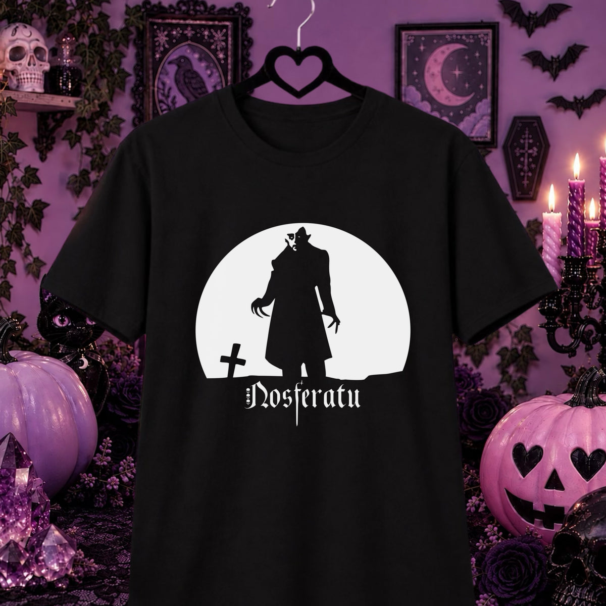A black short-sleeve t-shirt on a hanger, featuring a white circular graphic of a vampire silhouette with the gothic text 'Nosferatu' below it. The shirt is displayed against a dark, spooky Halloween-themed background with purple pumpkins, candles, and framed moon artwork.