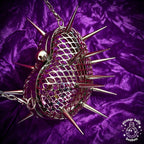 A metallic silver spiked heart-shaped bag with a mesh pattern, resting on a purple velvet fabric background. The bag features sharp metal spikes and a chain strap, with a 'Strange Dollz Boudoir' logo in the bottom right corner.