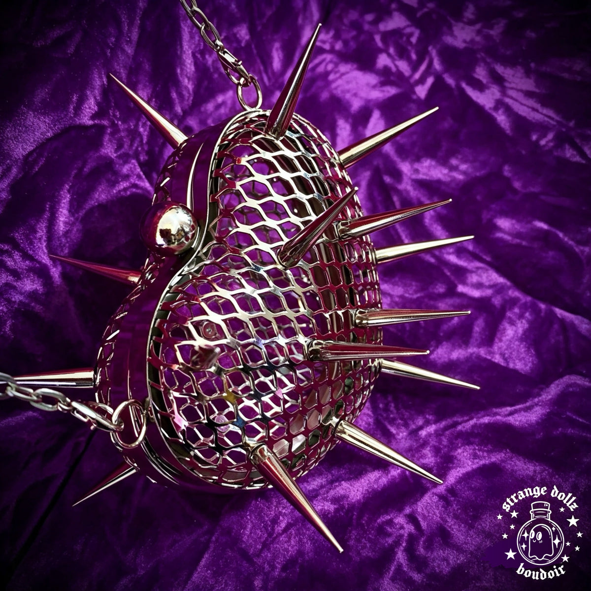 A metallic silver spiked heart-shaped bag with a mesh pattern, resting on a purple velvet fabric background. The bag features sharp metal spikes and a chain strap, with a 'Strange Dollz Boudoir' logo in the bottom right corner.