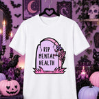 A white short-sleeve t-shirt on a black hanger, featuring a 'RIP MENTAL HEALTH' tombstone graphic in pink and black. The shirt is displayed against a dark purple Halloween-themed background with pumpkins, candles, and a black cat.