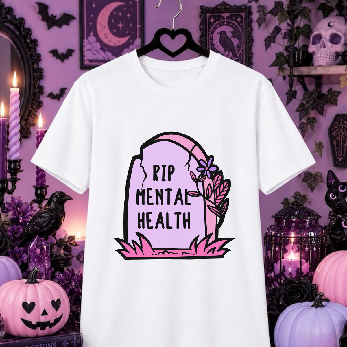 A white short-sleeve t-shirt on a black hanger, featuring a 'RIP MENTAL HEALTH' tombstone graphic in pink and black. The shirt is displayed against a dark purple Halloween-themed background with pumpkins, candles, and a black cat.