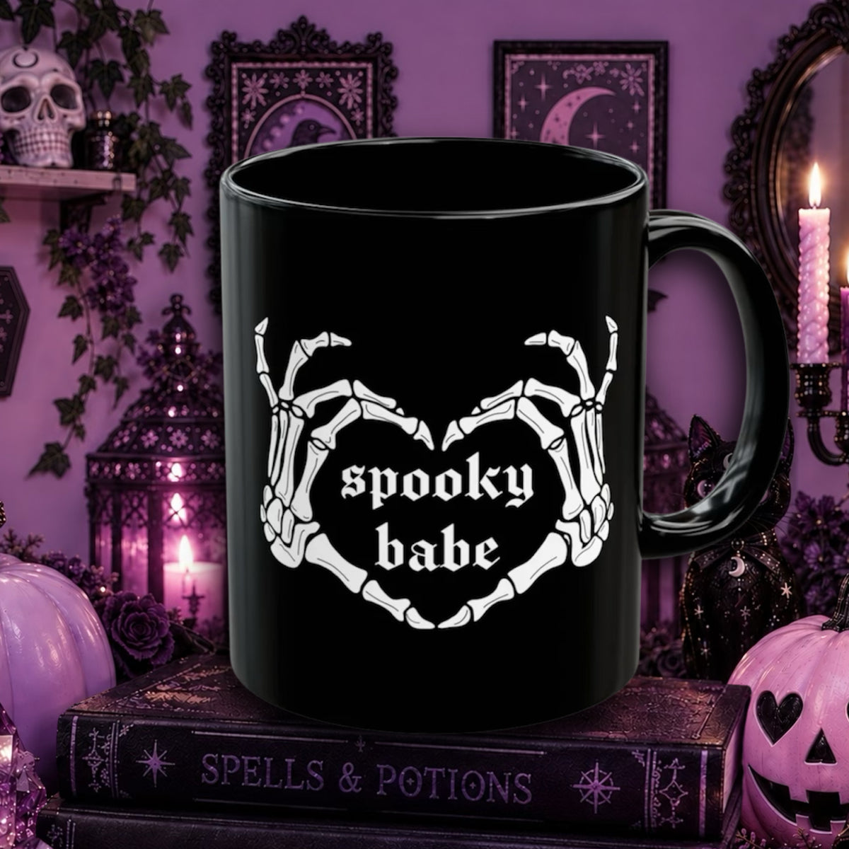 A black ceramic mug with a white skeleton hand graphic forming a heart shape around the words 'spooky babe' in gothic lettering. The mug is styled on a book surrounded by Halloween decorations, including candles, pumpkins, and a skull, against a purple background.