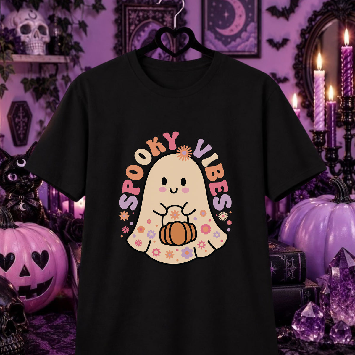 A black short-sleeve graphic t-shirt hanging on a black hanger against a purple Halloween-themed background. The shirt features a cute cartoon ghost holding a pumpkin with the text 'SPOOKY VIBES' in pastel colors.