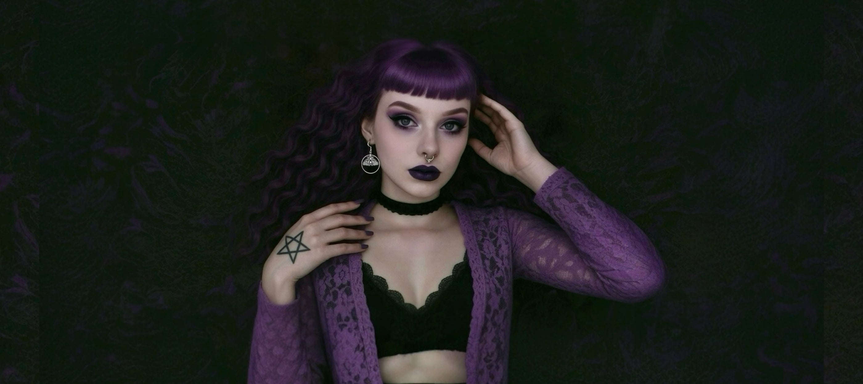 Woman with purple hair and dark makeup wearing a black top and purple cardigan against a dark background.