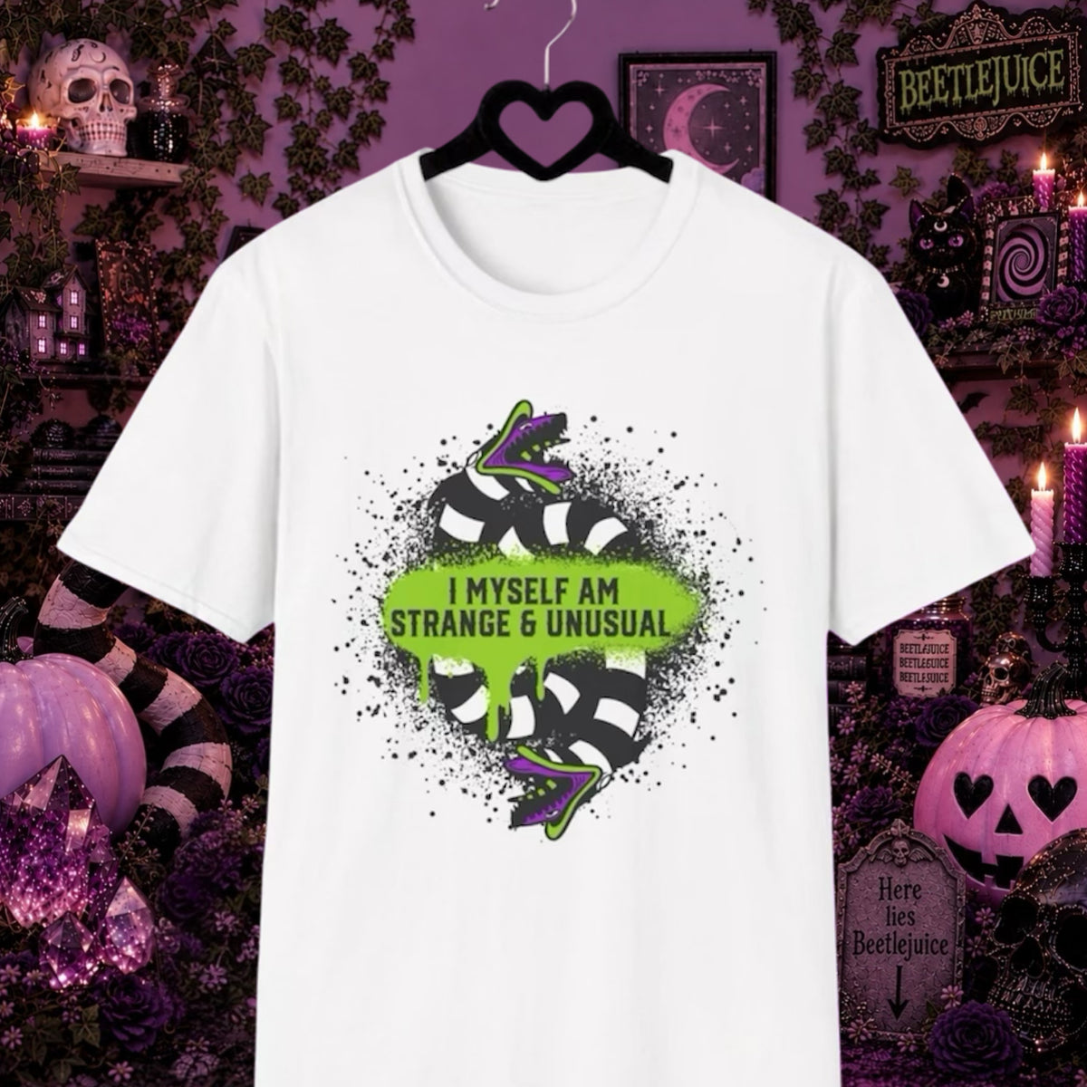 A white Beetlejuice Sandworm short-sleeve t-shirt hanging on a black hanger, featuring a graphic of a black and white striped snake with purple and green accents around a green heart shape with the text 'I MYSELF AM STRANGE & UNUSUAL'. The background is a dark, spooky Halloween-themed room with pumpkins, candles, and purple walls.